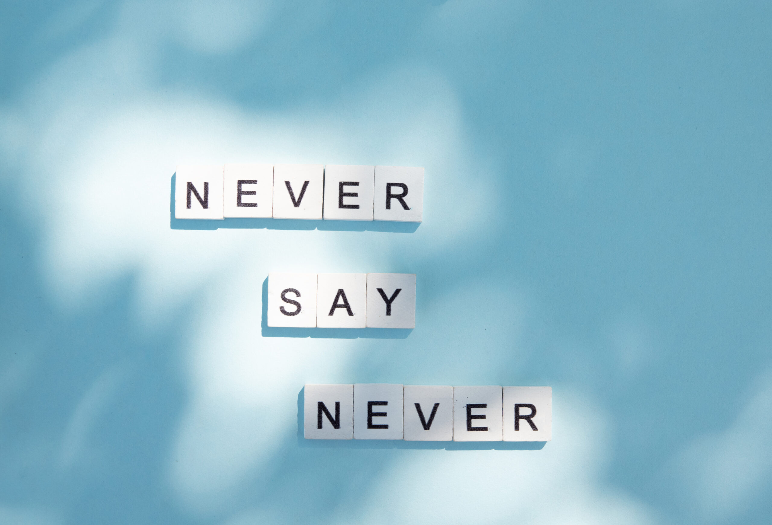 White letter tiles spelling Never Say Never on blue background representing resilience morse code jewelry by CeeBee